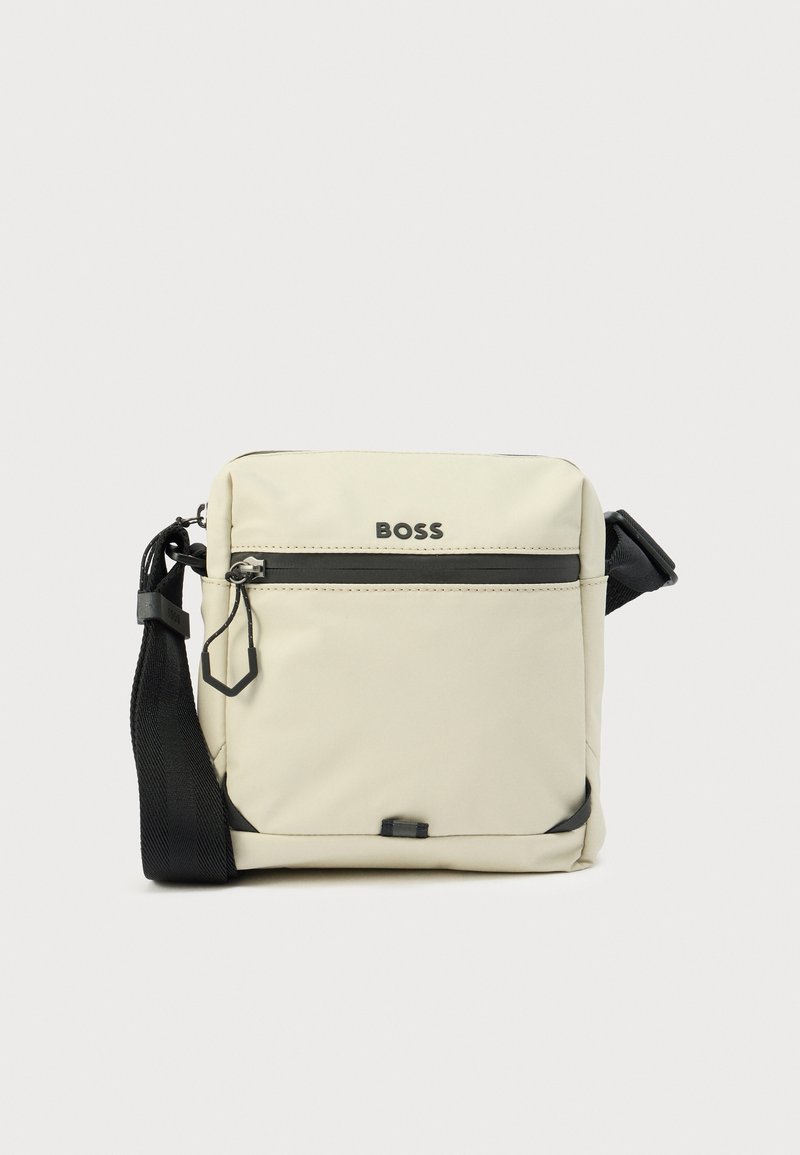 Beige Umhängetasche aus strapazierfähigem Stoff, mit einer vorderen Reißverschlusstasche, verstellbarem Riemen und schwarzen Akzenten. Logo auf der Vorderseite angebracht.