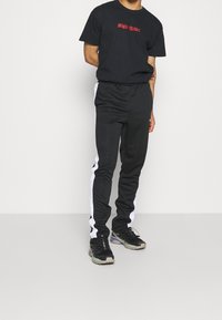 Svart t-shirt med röd text, kombinerad med svarta träningsoverallsbyxor med vita sidostripes. Bärs med svarta sportskor.