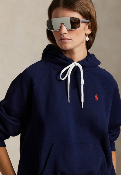 Γυναίκα που φοράει αντανακλαστικά, μεγάλα γυαλιά ηλίου, μια μπλε σκούρα φούτερ με άσπρα κορδόνια και ένα μικρό κόκκινο λογότυπο στο στήθος, με σκουλαρίκια κοχύλια.