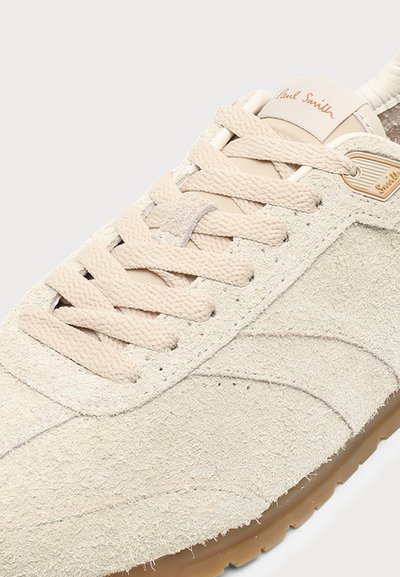 Gros plan d'une basket beige en daim avec des lacets texturés, une languette lisse et une semelle en gomme, portant la marque Paul Smith sur la languette et le côté.