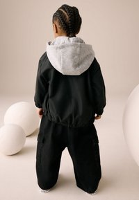 Veste noire avec une capuche grise, associée à un pantalon cargo noir surdimensionné. Tissu lisse avec des poignets élastiques. La scène présente des formes rondes blanches.