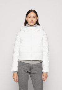 Hvid cropped pufferjakke med hætte, der har vandret quiltet design og lynlås foran. Båret over en lysegrå sweater og jeans.