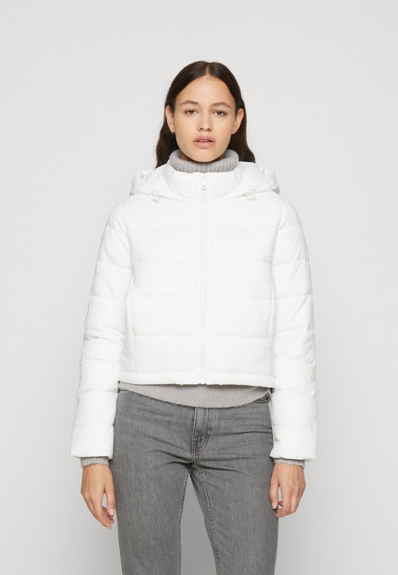 Hvid cropped pufferjakke med hætte, der har vandret quiltet design og lynlås foran. Båret over en lysegrå sweater og jeans.