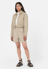 Zastrihnutá béžová bunda s rovným golierom, doplnená o béžové šortky s vysokým pásom. Čierne sandále na opätku dopĺňajú celý outfit.