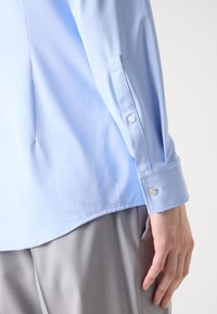 Personne portant une chemise bleu clair à manches longues avec poignets boutonnés et un pantalon gris clair, main reposant sur le côté.
