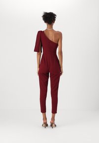 Combinaison bordeaux à une manche longue et dos nu. Design ajusté avec jambes fuselées et fermeture éclair au dos. Texture de tissu lisse.