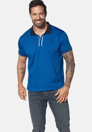 Blue polo shirt made of cotton, featuring a black and white collar, short sleeves, and a small logo on the chest. Worn with dark jeans.