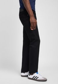 Homme portant un pantalon noir et des baskets blanches avec des bandes noires, debout devant un fond gris uni.