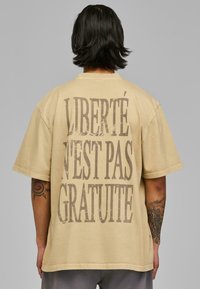Tricou oversized bej cu mâneci scurte, având imprimat îndrăzneț și uzat textul "LIBERTÉ N'EST PAS GRATUITE" pe spate. Material din bumbac.