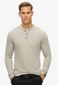 Lichtgrijs langvendig henley shirt met een ronde hals, drie donkere knopen en een gestructureerde stof. Gedragen over een wit shirt.
