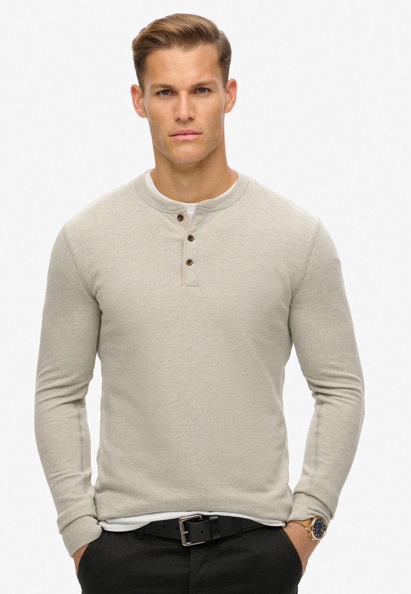 Lichtgrijs langvendig henley shirt met een ronde hals, drie donkere knopen en een gestructureerde stof. Gedragen over een wit shirt.
