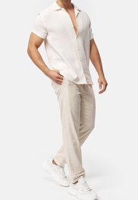 Kurzärmliges, hellbeiges Hemd mit Knöpfen und strukturierter Musterung, kombiniert mit hellbeigen Hosen und weißen Sneakers, was ein lässiges Outfit ergibt.