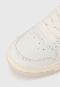 Vita lädersneakers med texturerad ovandel, krämfärgade snören, perforerad tåbox och beige gummisula. Diskret stickningsdetalj längs kanterna.