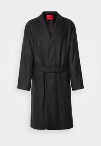 Manteau en laine gris foncé longueur genou avec revers crantés, trois boutons frontaux, ceinture à la taille, deux grandes poches frontales, et étiquette rouge HUGO à l'intérieur.