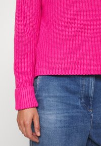 Maglione aderente rosa a costine con maniche larghe, abbinato a jeans blu. Il tessuto testurizzato è evidente nel design.