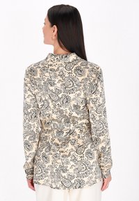 Koszula z długim rękawem w beżowym kolorze z czarnym wzorem w paisley i kwiatowym, z kołnierzykiem i mankietami zapinanymi na guziki. Widok z tyłu pokazuje wcięcie w talii.