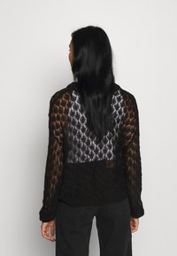 Kvinna med långt svart hår som bär en svart spetsstickad tröja och svarta byxor, vänd bort mot en enfärgad grå bakgrund.