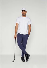 Valge lühikeste varrukatega T-särk, tumesinised dressipüksid, mustad kingad. Mees hoiab golfikeppi. Lihtne disain, vaba lõige ja ühetoonilised värvid.