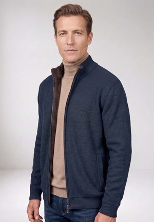 Mann trägt marineblauen Reißverschluss-Pullover über beigem Rollkragenpullover, steht vor einfarbig hellem Hintergrund und schaut geradeaus.