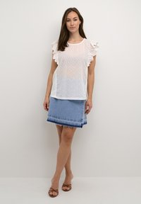 Blusa blanca de ojo de perdiz con mangas rizadas, combinada con una falda de mezclilla azul con dobladillo deshilachado y una franja de acento azul oscuro.