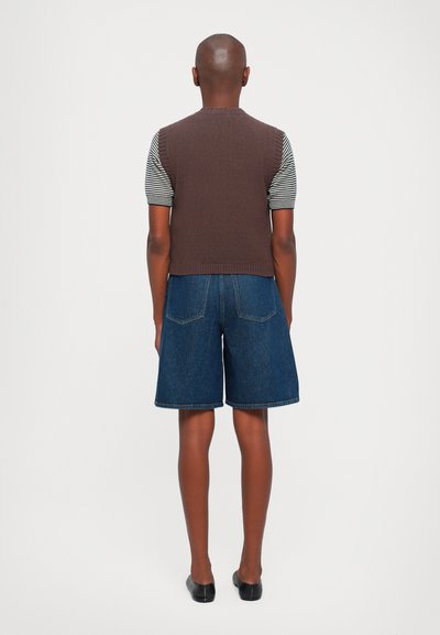Gilet marrone senza maniche sopra una maglietta a righe a maniche corte, abbinato a pantaloni denim. Tessuto liscio e design aderente con dettagli sulla schiena.