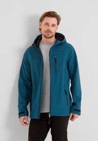 Teal softshell jacket with a black hood, zipper pockets, and contrasting black accents. Worn over a gray t-shirt, featuring a front zip closure.