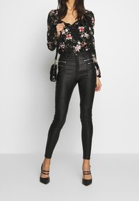 Pantalones ajustados de imitación cuero negro con bolsillos con cremallera. La parte superior presenta un patrón floral sobre un fondo negro, con un escote con volantes.