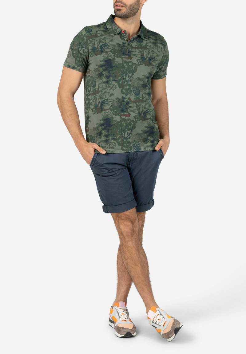 Green patterned polo shirt with a collar and two-button placket, paired with navy shorts. Model wears multicolored athletic sneakers.