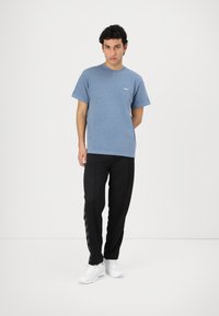 Giovane uomo in piedi che indossa una t-shirt blu a maniche corte, pantaloni neri con strisce laterali e scarpe da ginnastica bianche su uno sfondo bianco semplice.