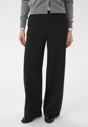 Persoană purtând pantaloni negri largi, cardigan gri și pantofi negri, stând în fața unui fundal simplu.