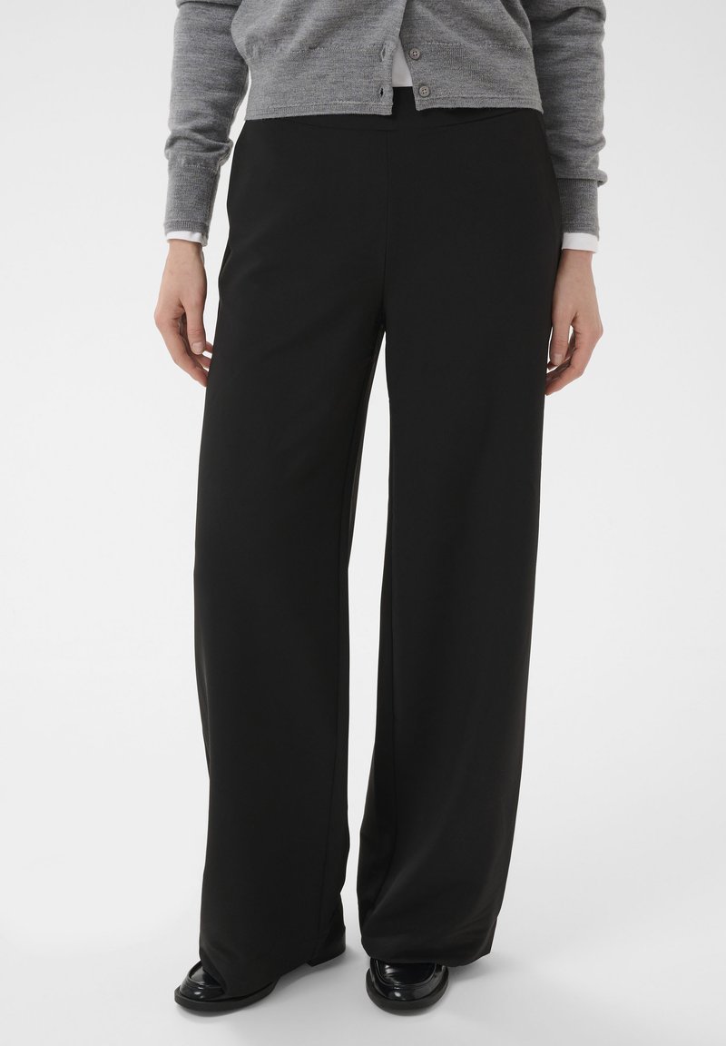 Personne portant un pantalon noir à jambes larges, un cardigan gris et des chaussures noires, debout devant un fond uni.