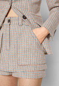 Blazer à motif pied-de-poule multicolore et shorts taille haute assortis, main dans la poche et boutons noirs sur la veste et les shorts.