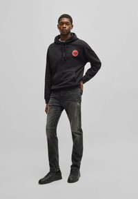 Felpa nera con logo rosso, tasca frontale a marsupio; abbinata a jeans grigi strappati e sneakers nere. Design semplice e casual.