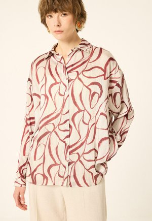 Femme portant un chemisier ample crème avec des lignes abstraites rouges et un pantalon beige, debout avec une main dans la poche devant un fond uni.
