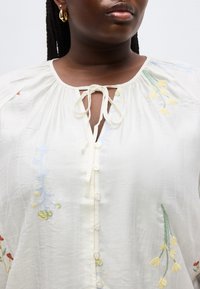 Femme portant un chemisier blanc à boutons sur le devant avec une broderie florale pastel, un col noué et des créoles en or.