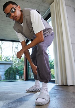 Gilet gris sans manches sur une chemise blanche, associé à un pantalon gris ample et des chaussures blanches. Environnement intérieur avec de grandes fenêtres et un sol en béton.
