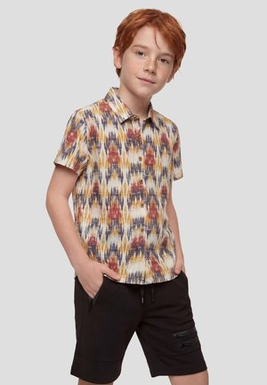 Niño pelirrojo con camisa de manga corta estampada multicolor y pantalones cortos negros con las manos en los bolsillos, de pie contra un fondo liso.