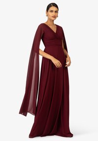 Abito da sera bordeaux con corpetto aderente, dettagli plissettati e maniche trasparenti e fluide. Realizzato in tessuto leggero, ha una forma lunga ed elegante.