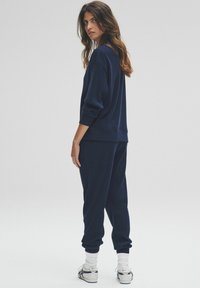 Mujer de pie de perfil que lleva un suéter de canalé azul marino y pantalones con calcetas blancas y zapatillas deportivas, frente a un fondo claro y liso.