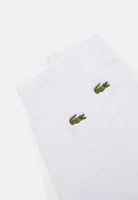 Calcetines de algodón blancos con puños acanalados, que presentan un pequeño logo de cocodrilo bordado en verde en el lado exterior. Textura suave, altura estándar de tobillo.