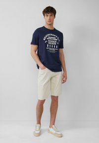 Navy t-shirt met witte grafische print, gecombineerd met beige shorts. Model draagt lichtkleurige sneakers. Minimalistisch ontwerp, casual pasvorm.