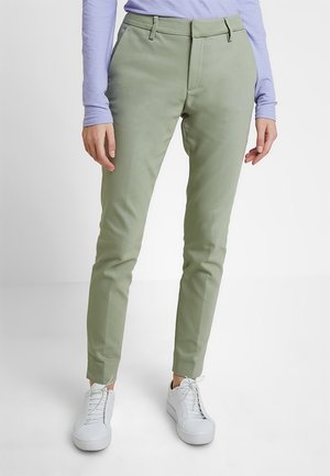 Persoon draagt lichtgroene slim-fit broek, witte sneakers en een lichtpaarse longsleeve, staand tegen een effen witte achtergrond.