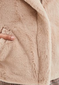 Lichtbeige nepbontjas met een zachte textuur, brede kraag en zijzakken, gedeeltelijk bedekt door een hand.