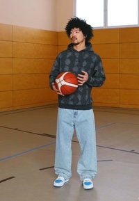 Čierny vzorovaný mikina, svetlo modré voľné džínsy a tenisky s modrými akcentmi. Drží basketbal v telocvični s drevenými stenami.