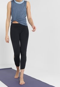 Blå ärmlös knuten topp med rund halsringning, i kombination med svarta caprileggings med utskärningar på sidorna. Står på en yogamatta.