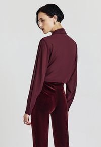 Blusa di seta borgogna con colletto, maniche lunghe e una texture liscia, abbinata a pantaloni in velluto borgogna a vita alta.