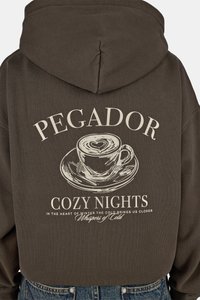 Barna kapucnis pulóver kávéscsészét és alátétet ábrázoló grafikával, a "PEGADOR COZY NIGHTS" felirattal világos bézs színben, puha tapintással.