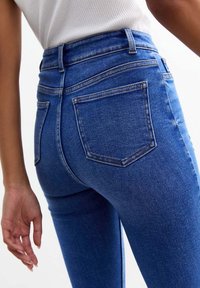 Acercamiento de una persona que lleva vaqueros de mezclilla azul de cintura alta y una camiseta sin mangas blanca de canalé, mostrando los bolsillos traseros y parte de la mano.