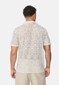 Chemisette à manches courtes en blanc cassé avec des motifs complexes ; tissu léger et respirant affichant un design en crochet détaillé.