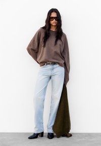 Brauner oversized Sweatshirt, hellblaue gerade Jeans, klobige schwarze Stiefel mit Absatz und ein dunkelgrüner strukturierter Pullover über den Arm drapiert.